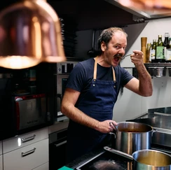 Mann mit dunklem T-Shirt und Schürze steht in moderner Küche und kostet mit Löffel aus großem Topf