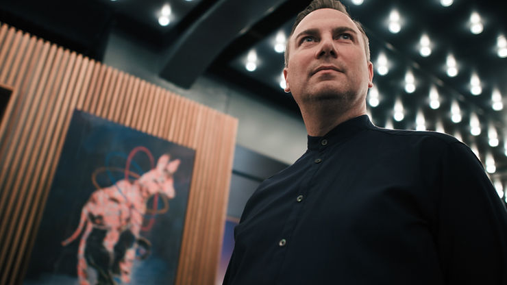 Tim Raue in schwarzem Hemd steht vor einer Holzvertäfelung mit einem Bild, das ein rosa Tier zeigt