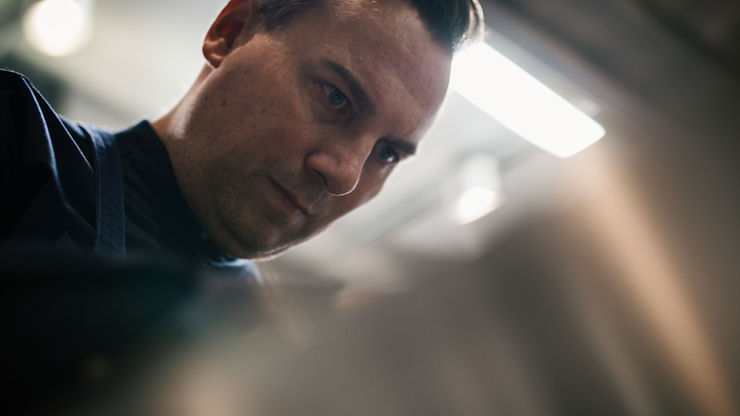 Tim Raue in dunkler Arbeitskleidung mit Schürze in einer Werkstatt oder Küche vor unscharfem Hintergrund