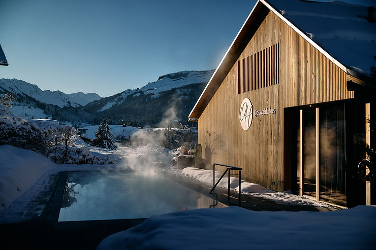 Holzgebäude neben einem beheizten Außenpool mit Dampfschwaden in einer verschneiten Berglandschaft