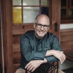 Mann mit dunklem Hemd sitzt mit verschränkten Armen vor einer Holzwand mit Fenster