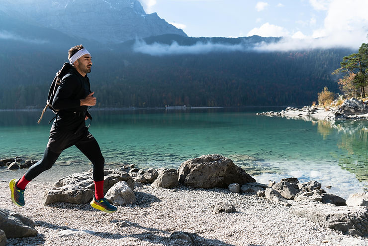 Savas Coban in Sportkleidung läuft am Ufer eines Bergsees mit Bergen und Wolken im Hintergrund
