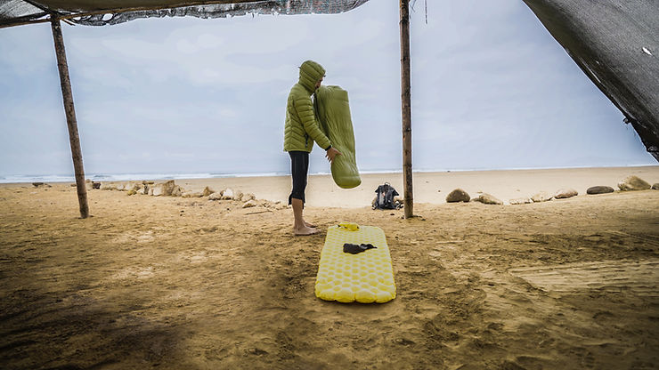 Savas Coban mit Kapuzenjacke steht barfuß auf Sand unter einem Zelt und hält eine aufgerollte Isomatte über einer gelben Luftmatratze