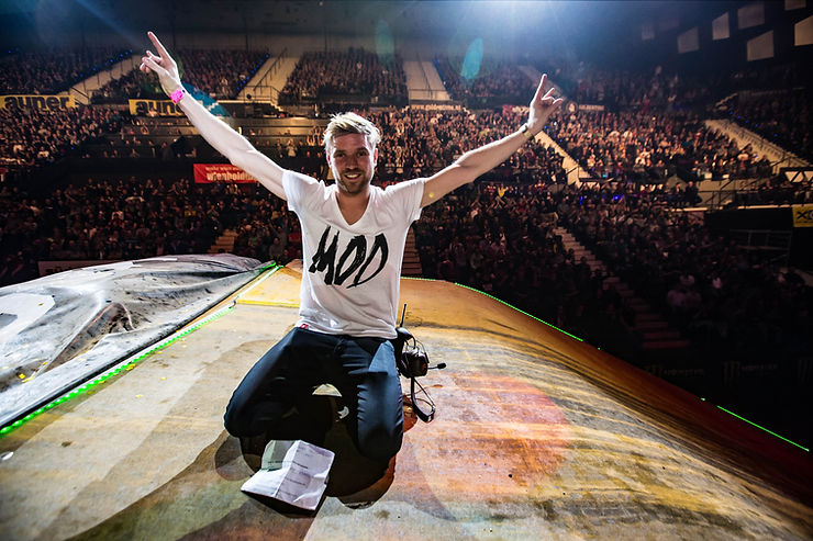 Georg Fechter kniet auf einer Bühne vor einem großen Publikum und hebt beide Arme mit ausgestreckten Zeigefingern nach oben