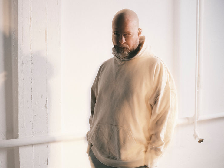 Curse mit Glatze trägt einen hellen Kapuzenpullover und steht vor einer weißen Wand mit sichtbaren Rohren
