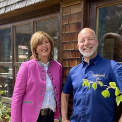 Zwei Personen stehen nebeneinander vor einem Holzhaus mit Fenstern, die linke trägt eine pinke Jacke und die rechte ein blaues Hemd