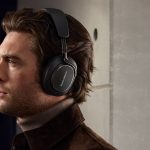 Person mit braunen Haaren trägt schwarze Kopfhörer und braunen Mantel vor einer grauen Wand