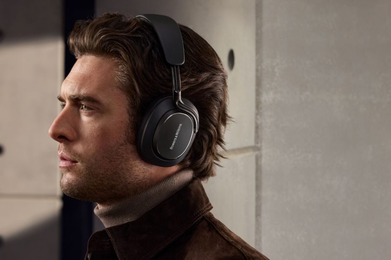 Person mit braunen Haaren trägt schwarze Kopfhörer und braunen Mantel vor einer grauen Wand