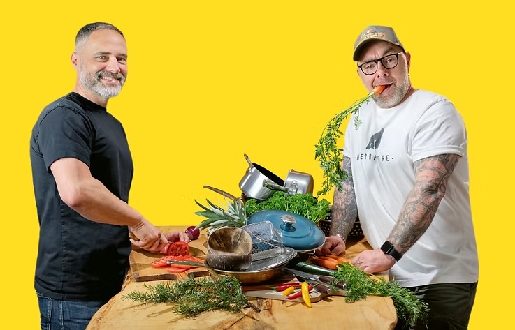 Alexander Flohr und Kai Lüftner stehen an einem Holztisch mit verschiedenen Küchenutensilien, Gemüse und Kräutern vor gelbem Hintergrund