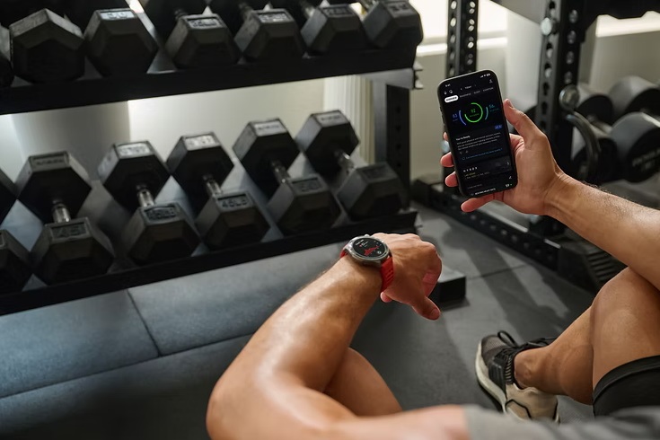 Person sitzt im Fitnessstudio vor einer Hantelablage und hält ein Smartphone mit Fitness-App in der Hand, am Handgelenk eine Sportuhr