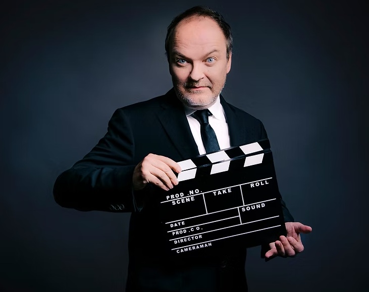 Jens Wawrczeck im schwarzen Anzug mit weißem Hemd und schwarzer Krawatte hält eine Filmklappe vor dunklem Hintergrund.