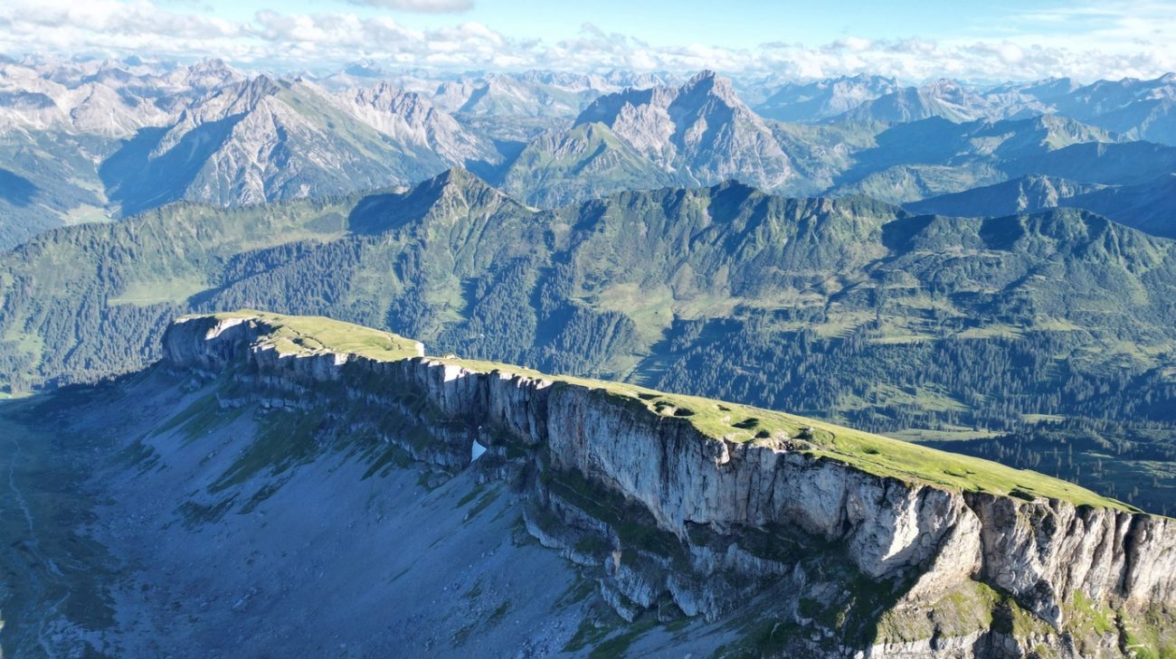 Luftaufnahme eines langgezogenen Bergkamms mit steilen Felswänden und grasbewachsenen Flächen vor einer Berglandschaft mit bewaldeten Hängen und Gipfeln unter blauem Himmel mit Wolken