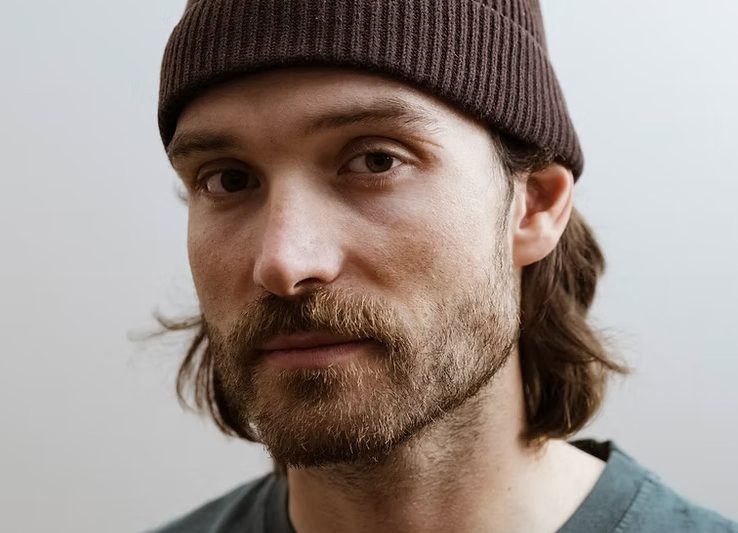 Rick Zabel mit brauner Strickmütze und dunkelgrünem T-Shirt vor hellem Hintergrund