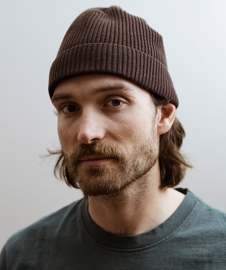 Rick Zabel mit brauner Strickmütze und dunkelgrünem T-Shirt vor hellem Hintergrund