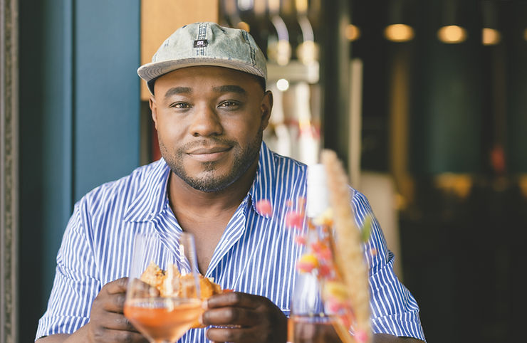 Nelson Müller mit gestreiftem Hemd und Baseballkappe sitzt an einem Tisch mit Getränken und hält ein Sandwich in der Hand