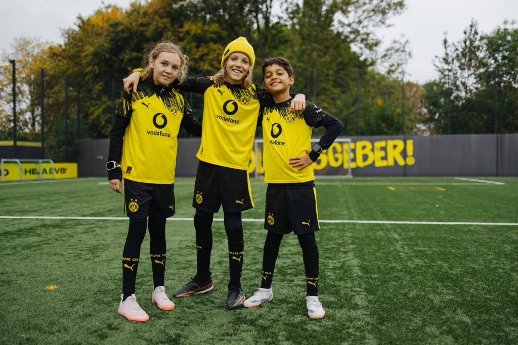 Drei Kinder in BVB-Trikots mit Xplora X6Play BVB-Edition auf dem Fußballplatz